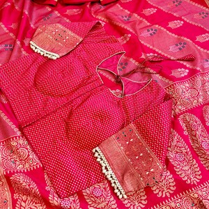 May include: A red and gold patterned silk saree with a matching red blouse featuring white polka dots. The blouse has a keyhole neckline and decorative gold trim.