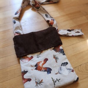 May include: A rectangular fabric crossbody bag featuring a light gray background with a repeating pattern of colorful roosters and small floral sprigs. The bag has a dark brown rectangular flap closure and a matching long fabric shoulder strap.