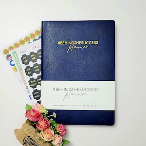 Peut inclure: Un agenda bleu marine avec le texte "#BEHINDTHESUCCESS planner" en lettres dorées. L'agenda est non daté et a une mise en page hebdomadaire. Il mesure 17,5 cm x 22 cm et 120 g/m².