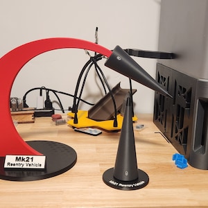 Op de afbeelding: Een 3D-geprint rood en zwart model van een raket, met het label "Mk.21 Reentry Vehicle". Het model staat op een zwarte basis en staat op een houten tafel.
