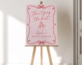 May include: A rectangular light pink sign with a red decorative border featuring bows. The text reads: She's tying the knot, WELCOME TO Angelica's BRIDAL SHOWER, SEPTEMBER 16, 2023. A red line drawing of two clinking champagne glasses is centered.