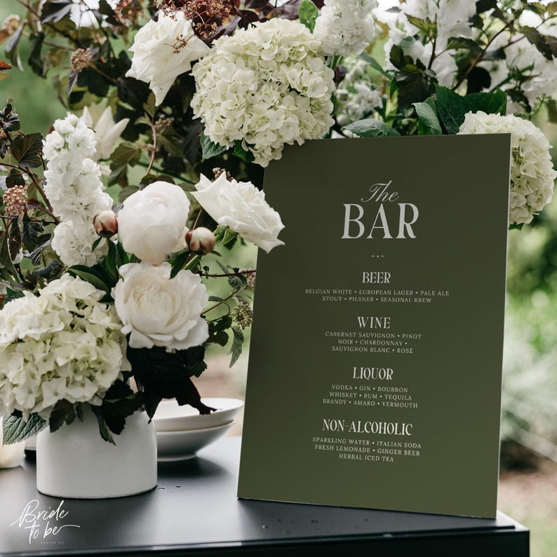 May include: A dark green bar menu with white text, listing beer, wine, liquor, and non-alcoholic beverages. The menu is set against a backdrop of white flowers and greenery, perfect for a wedding or event.