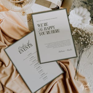 May include: Two wedding celebration signs with green borders and text. One says "Order of Events" and lists times. The other says "We're so happy you're here." Both are on a beige cloth with flowers.