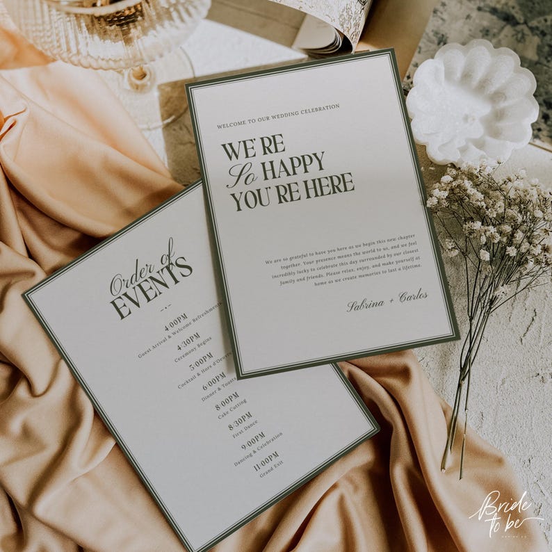 May include: Two wedding celebration signs with green borders and text. One says "Order of Events" and lists times. The other says "We're so happy you're here." Both are on a beige cloth with flowers.
