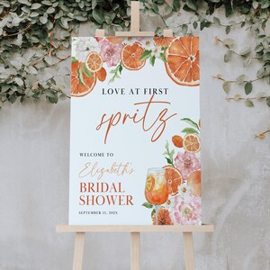 May include: A bridal shower welcome sign with a watercolor illustration of orange slices, flowers, and a glass of Aperol Spritz. The text reads "Love at First Spritz, Welcome to Elizabeth's Bridal Shower, September 13, 202X".