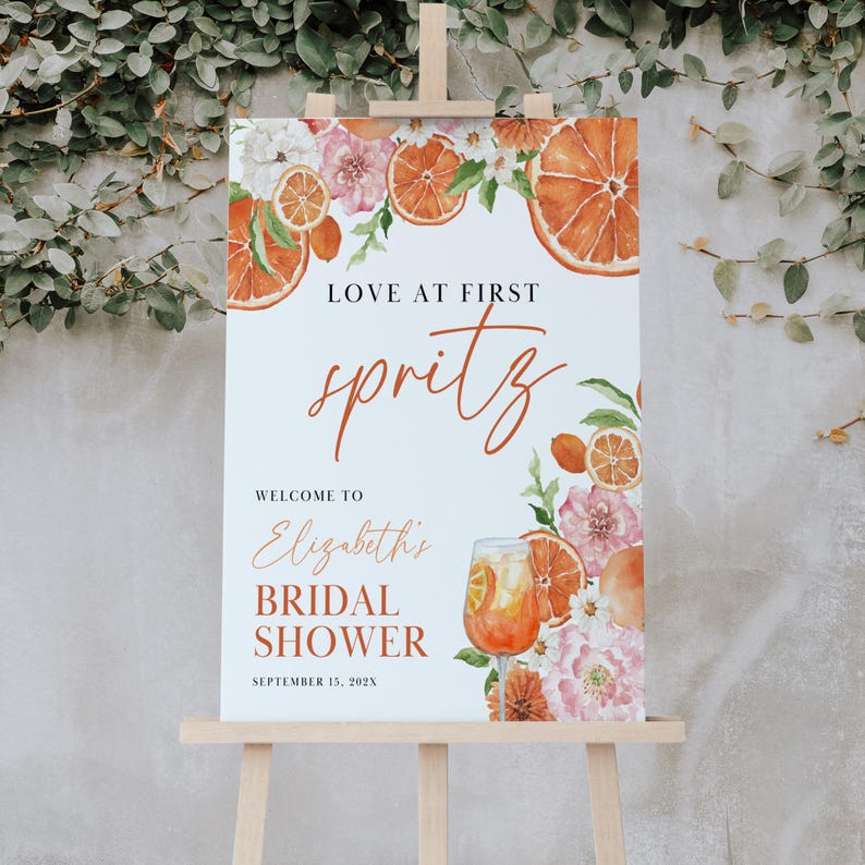 May include: A bridal shower welcome sign with a watercolor illustration of orange slices, flowers, and a glass of Aperol Spritz. The text reads "Love at First Spritz, Welcome to Elizabeth's Bridal Shower, September 13, 202X".