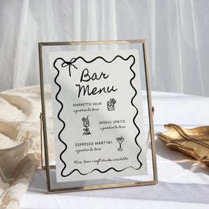 May include: A gold-framed sign with a white background and black text lists a bar menu with three drink options: Amaretto Sour, Aperol Spritz, and Espresso Martini. The sign also states that wine, beer, and soft drinks are available.
