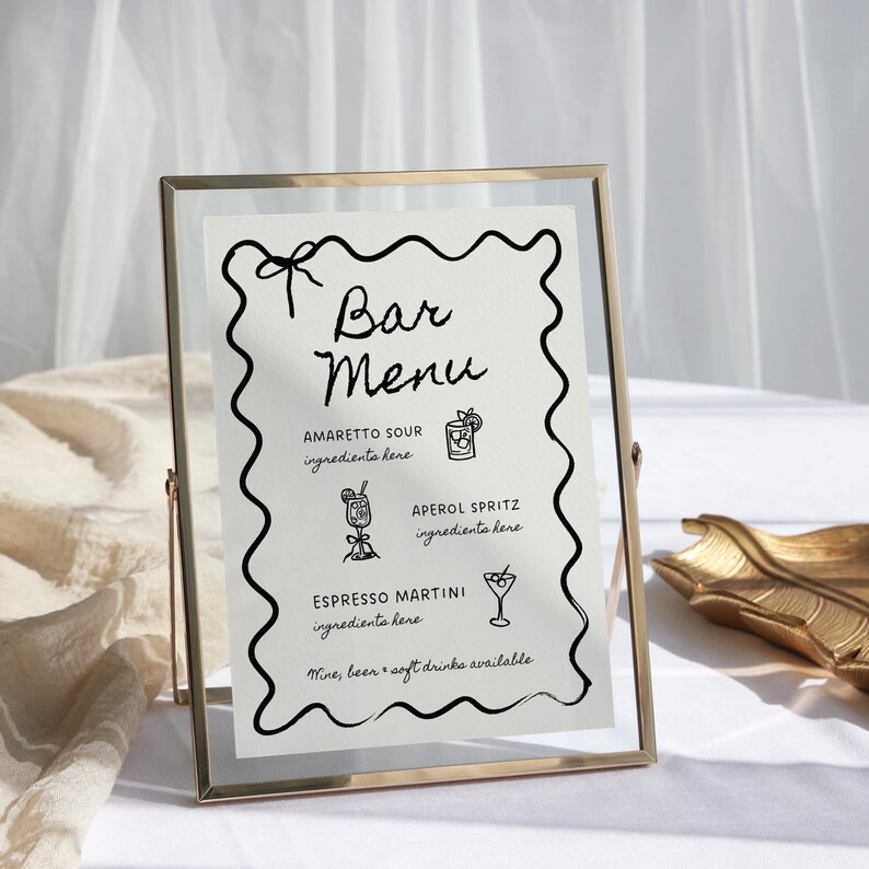 May include: A gold-framed sign with a white background and black text lists a bar menu with three drink options: Amaretto Sour, Aperol Spritz, and Espresso Martini. The sign also states that wine, beer, and soft drinks are available.