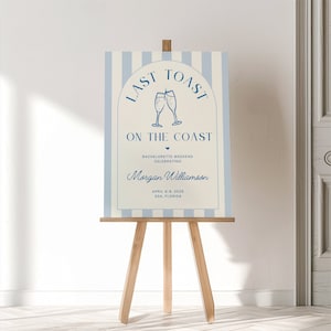 May include: A white and blue striped sign with the text "Last Toast on the Coast" in a blue arch. The sign also includes a blue illustration of two champagne glasses, the text "Bachelorette Weekend Celebrating", a name, and the date and location of the event.