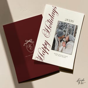 Może przedstawiać: Dwie kartki świąteczne. Jedna jest bordowa z białym logo i tekstem. Druga jest w kolorze złamanej bieli ze słowami "Happy Holidays" w bordowym skrypcie. Zdjęcie całującej się pary znajduje się na środku, nad nim rok 2026.