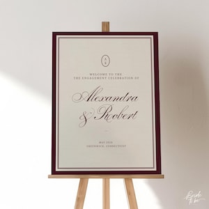 May include: A framed engagement celebration sign on a wooden easel. The sign has a cream-colored background with burgundy borders and text. The text reads "Welcome to the Engagement Celebration of Alexandra & Robert, May 2026, Greenwich, Connecticut."