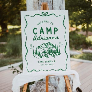 Può includere: Un cartello bianco con testo e illustrazioni verdi, esposto su un cavalletto di legno. Il cartello recita "WELCOME TO CAMP Adrianna" sopra un grafico di montagna e cabina. Sotto, c'è scritto "LAKE TAHOE, CA JULY 11-13, 2025".