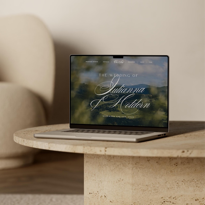 May include: A silver laptop displaying a wedding website. The screen shows the words "The Wedding of Julianna Holden" in elegant script over a blurred landscape. The laptop sits on a round, beige stone table.