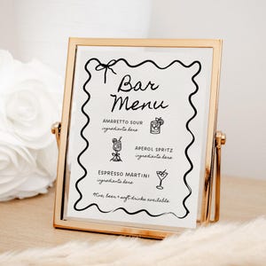 May include: A gold-framed sign with a black and white bar menu. The menu lists drinks like Amaretto Sour, Aperol Spritz, and Espresso Martini. The sign says "Bar Menu" at the top.