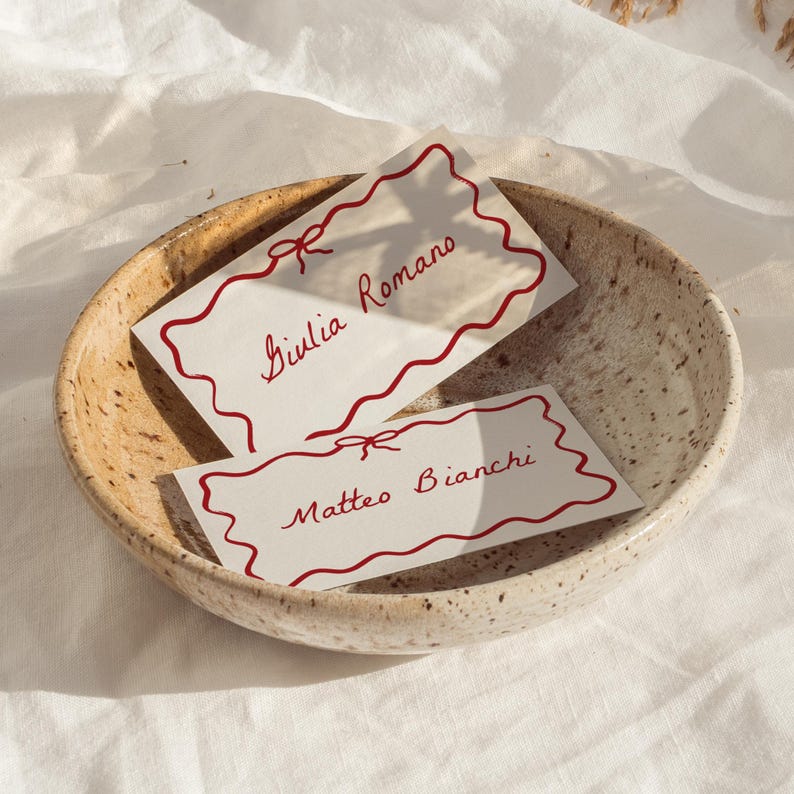 May include: Two white place cards with red wavy borders and red ribbon bows. The place cards are in a speckled brown bowl. The text on the place cards reads "Giulia Romano" and "Matteo Bianchi".