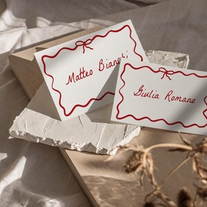 May include: Two white place cards with red scalloped borders and red ribbon bows. The text on the cards reads "Matteo Bianchi" and "Giulia Romano".