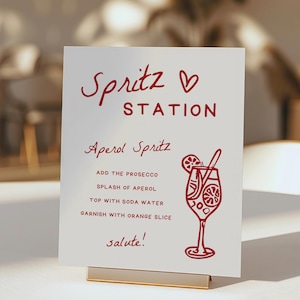 Könnte beinhalten: Ein weißes Schild mit rotem Text, auf dem "Spritz Station" steht und Anweisungen zur Zubereitung eines Aperol Spritz. Das Schild zeigt eine rote Illustration eines Glases Aperol Spritz mit einer Orangenscheibe und einem Strohhalm.