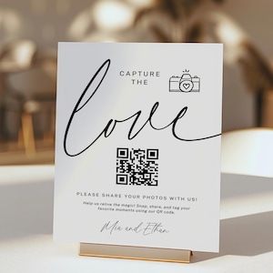 May include: White sign with black text that says "Capture the love" with a black and white camera icon with a heart inside. Below the text is a black and white QR code. Below the QR code is more black text that says "Please share your photos with us! Help us relive the magic! Snap, share, and tag your favourite moments using our QR code. Mia and Ethan"