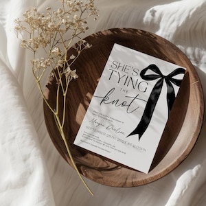 May include: A white bridal shower invitation with black text that reads "She's Tying the Knot" with a black satin bow. The invitation includes the date and time of the event, the bride-to-be's name, and the location.