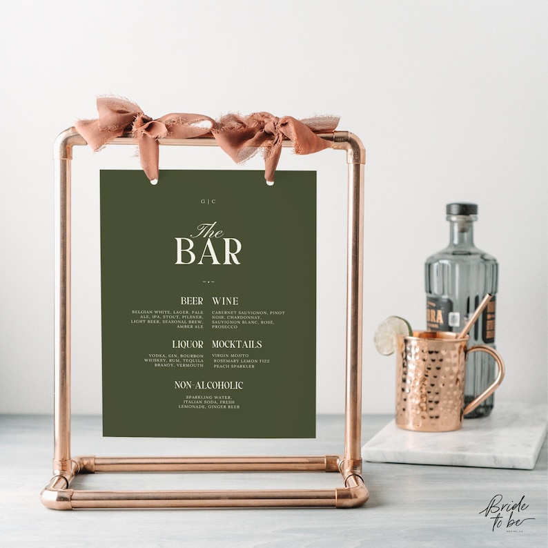 May include: A copper-colored metal bar sign with a dark green background and the words "The Bar" in white. The sign lists beer, wine, liquor, and mocktail options. A copper mug and a bottle of liquor are in the background.