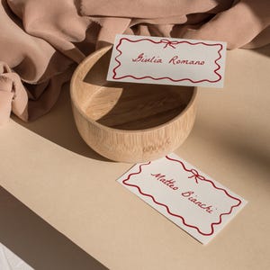 May include: Two white cards with red borders and red bows. The cards have the names "Giulia Romano" and "Matteo Bianchi" written on them. The cards are sitting on a light brown surface next to a wooden bowl.