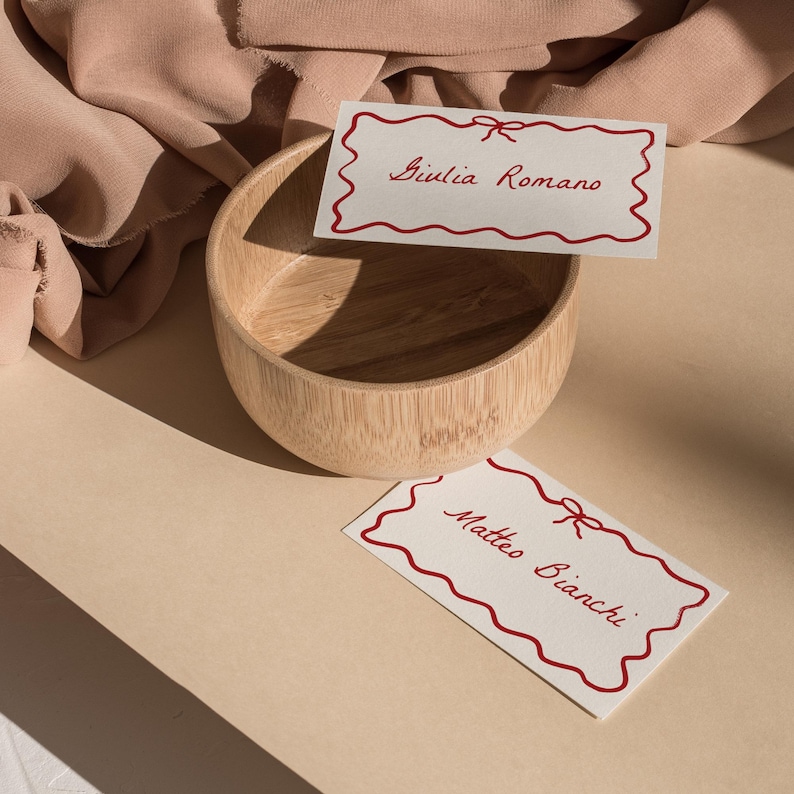 May include: Two white cards with red borders and red bows. The cards have the names "Giulia Romano" and "Matteo Bianchi" written on them. The cards are sitting on a light brown surface next to a wooden bowl.