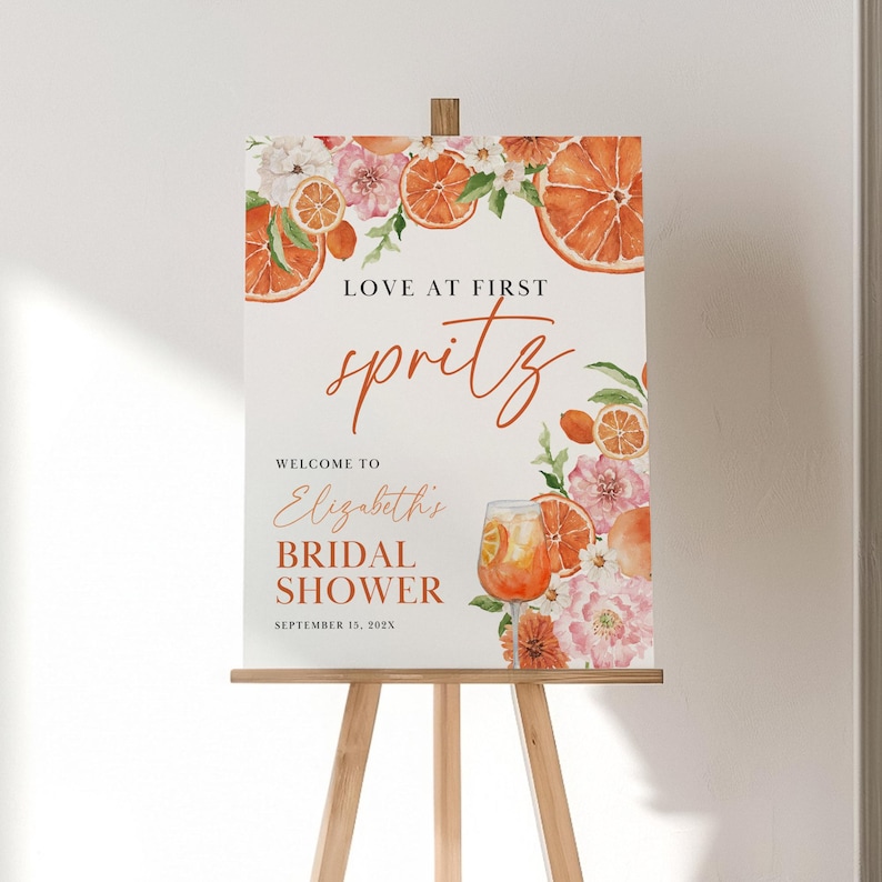 May include: Orange and floral bridal shower welcome sign with the text "Love at First Spritz" and "Welcome to Elizabeth's Bridal Shower September 15, 202X".