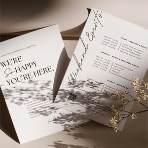 May include: A wedding invitation with a white background and black text. The invitation includes a welcome message, a weekend schedule of events, and a QR code. The text reads "We're so happy you're here." and "Weekend Events".