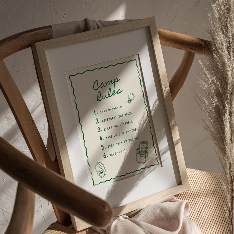 May include: A framed print with a white background and green text that reads "Camp Rules" with a list of rules for a camping trip. The rules include: Stay hydrated, Celebrate the bride, Relax and destress, Take lots of pictures, Stay cozy by the fire, and Have fun.