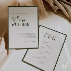 May include: Two wedding signs with green borders. One sign reads "WE'RE So HAPPY YOU'RE HERE" and the other lists the "Order of Events." The signs are on a white surface with a soft, neutral background.