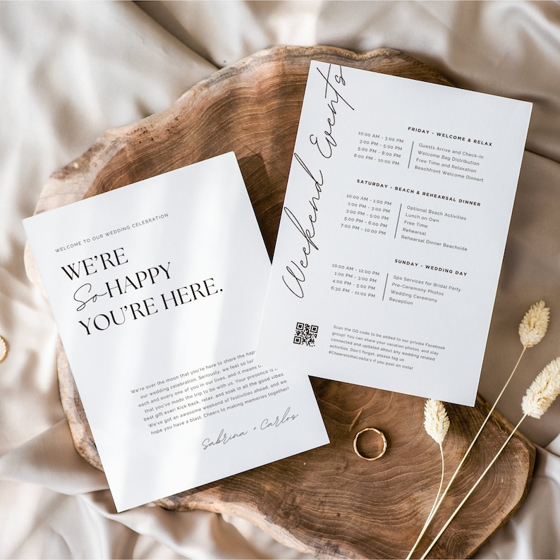 May include: A white wedding invitation with black text that reads "Welcome to our wedding celebration. We're so happy you're here." The invitation also includes a QR code and the names Sabrina and Carlos. The invitation is on a wooden surface with dried flowers and a gold ring.