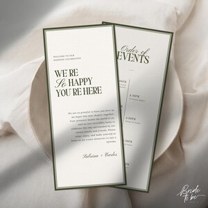 May include: Two wedding invitation cards with a green border and text. The first card reads "WE'RE So HAPPY YOU'RE HERE" and "WELCOME TO OUR WEDDING CELEBRATION". The second card reads "Order of EVENTS". The cards are on a white surface.