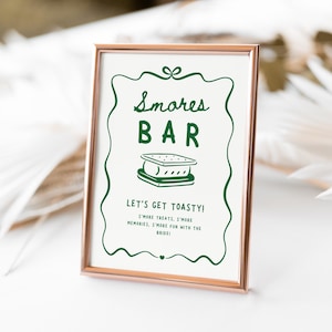 May include: A framed sign with a rose gold border. The sign has a white background with green text that reads "Smores BAR" and "LET'S GET TOASTY!". A graphic of a s'more is in the center. The sign is for a wedding or party.
