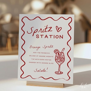 May include: A white sign with red text and a decorative border. The sign reads "Spritz Station" and includes instructions for an Orange Spritz cocktail. A gold-colored metal stand holds the sign.