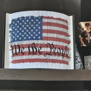 May include: A framed book art piece depicting the American flag. The flag design is created by folding the pages of the book, with red and white stripes and blue field with white stars. The words "We the People" are visible.