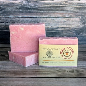 Op de afbeelding: Drie roze grapefruit zeepblokken. Eén zeepblok heeft een label met de tekst "Pink Grapefruit with Coconut Milk" en "Wild Acorn Artisan Soaps". Het nettogewicht is 130 g. De zeepblokken liggen op een houten oppervlak.