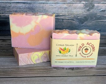 Op de afbeelding: Drie handgemaakte zeepblokken met een gemarmerd ontwerp in roze, oranje en gele tinten. Eén zeep is verpakt in een etiket met de tekst "Citrus Splash" en "Wild Acorn Artisan Soaps". Het nettogewicht is 130 g.