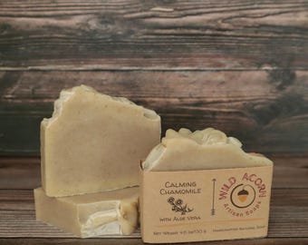 May include: Three bars of artisan soap with a light beige color, one partially cut. A small cardboard box displays "Calming Chamomile" and "Wild Acorn Artisan Soaps". The net weight is 4.6 oz/130 g. The background is a wooden surface.