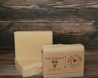 Może przedstawiać: Trzy ręcznie robione kostki mydła, z których jedna ma etykietę z napisem "Pure & Gentle With Goat Milk" i "Wild Acorn Artisan Soaps". Mydło jest w jasnobeżowym kolorze i waży 130 g.