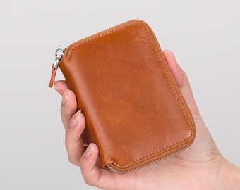 May include: A rectangular, brown leather wallet with a zipper closure. The wallet is held in a hand and has a smooth, textured surface. The zipper pull is a small, matching leather tab.