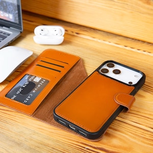 May include: A brown leather phone case with a wallet design, featuring card slots and a magnetic clasp. The case is open, revealing a credit card. A black and white phone is inside the case. A laptop, mouse, and earbuds are in the background.