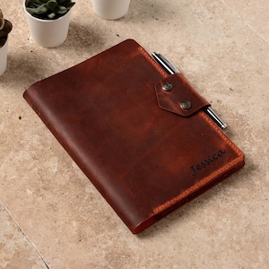 May include: A brown leather journal with a pen holder and a flap closure. The journal is personalized with the name "Jessica".