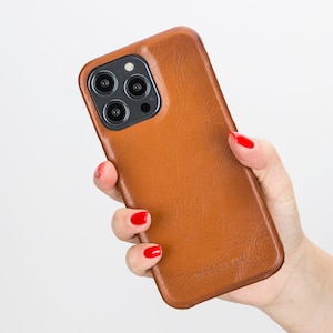 May include: A brown leather phone case with a black camera lens cutout. The case has a subtle texture and is held in a hand with red nail polish.