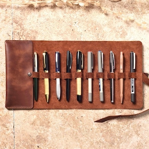 May include: A brown leather pen roll with 10 slots for pens and pencils. The roll is unrolled and the slots are filled with various writing instruments.