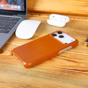 May include: A brown leather phone case rests on a wooden surface, next to a laptop, a white mouse, and a pair of white wireless earbuds. A patterned pen and a small gold coin are also visible.