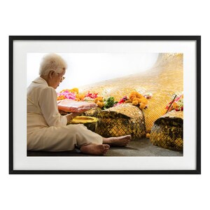 Peut inclure: Photographie encadrée d'une femme en vêtements clairs assise près d'une grande statue dorée. La scène comprend des offrandes florales et des feuilles d'or, suggérant un contexte religieux ou culturel.