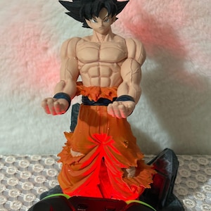 Op de afbeelding: Een zwart en oranje anime-figuur van een personage met stekelig zwart haar en een gescheurde oranje outfit. Het figuur houdt zijn handen voor zich uit. Het figuur wordt van onderen verlicht met rood licht. Een zwarte videogamecontroller bevindt zich op de voorgrond.