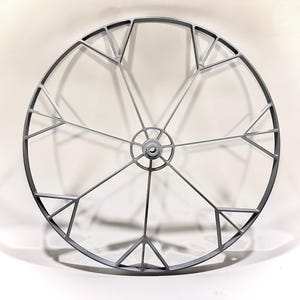 May include: A silver metal wheel with a geometric design. The wheel has a central hub and spokes that radiate outwards. The spokes are connected by triangular shapes.