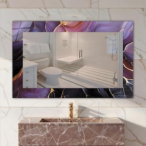 Puede incluir: Un espejo rectangular de baño con un borde decorativo con un diseño abstracto púrpura, negro y dorado. El espejo refleja un baño moderno con inodoro, ducha y lavabo blancos.