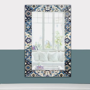 K&ouml;nnte beinhalten: Rechteckiger Spiegel mit einem dekorativen Rand mit blauem, wei&szlig;em und beigem Blumenmuster. Der Spiegel reflektiert ein helles Fenster mit wei&szlig;em Rahmen und einen Blick auf einen Raum mit wei&szlig;en Kissen und einem wei&szlig;en Sofa. Der Spiegel ist an einer zweifarbigen Wand befestigt.